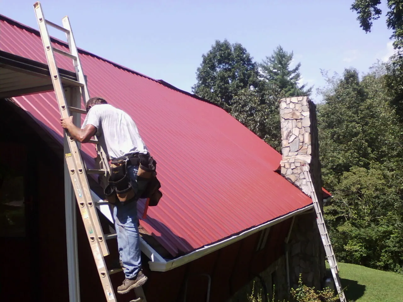 Roofer on a ladder beside a red metal roof and stone chimney — Soffit & Fascia Repair by Roofing Brothers in Woodstown